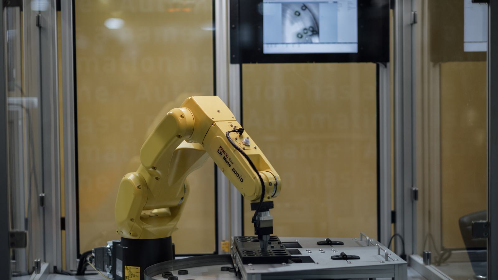 Industrial robot arm operating in an automated lab, with a vision-system monitor behind it